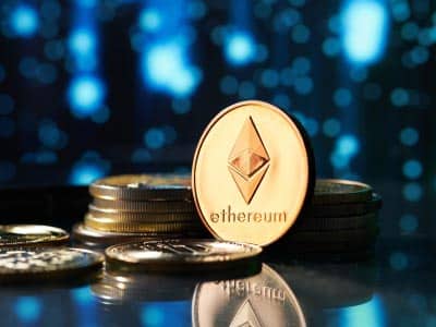 A close-up image of a photo-realistic golden Ethereum coin on a surface with other Ether coins against a blue blurry background